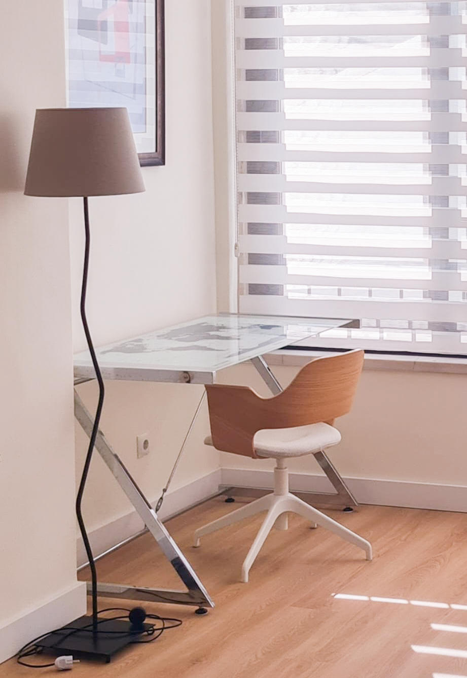 A  home office corner with a black floor lamp, a glass-top desk with metal legs, a wooden chair with a white cushion, a window with striped blinds, and a framed wall art.