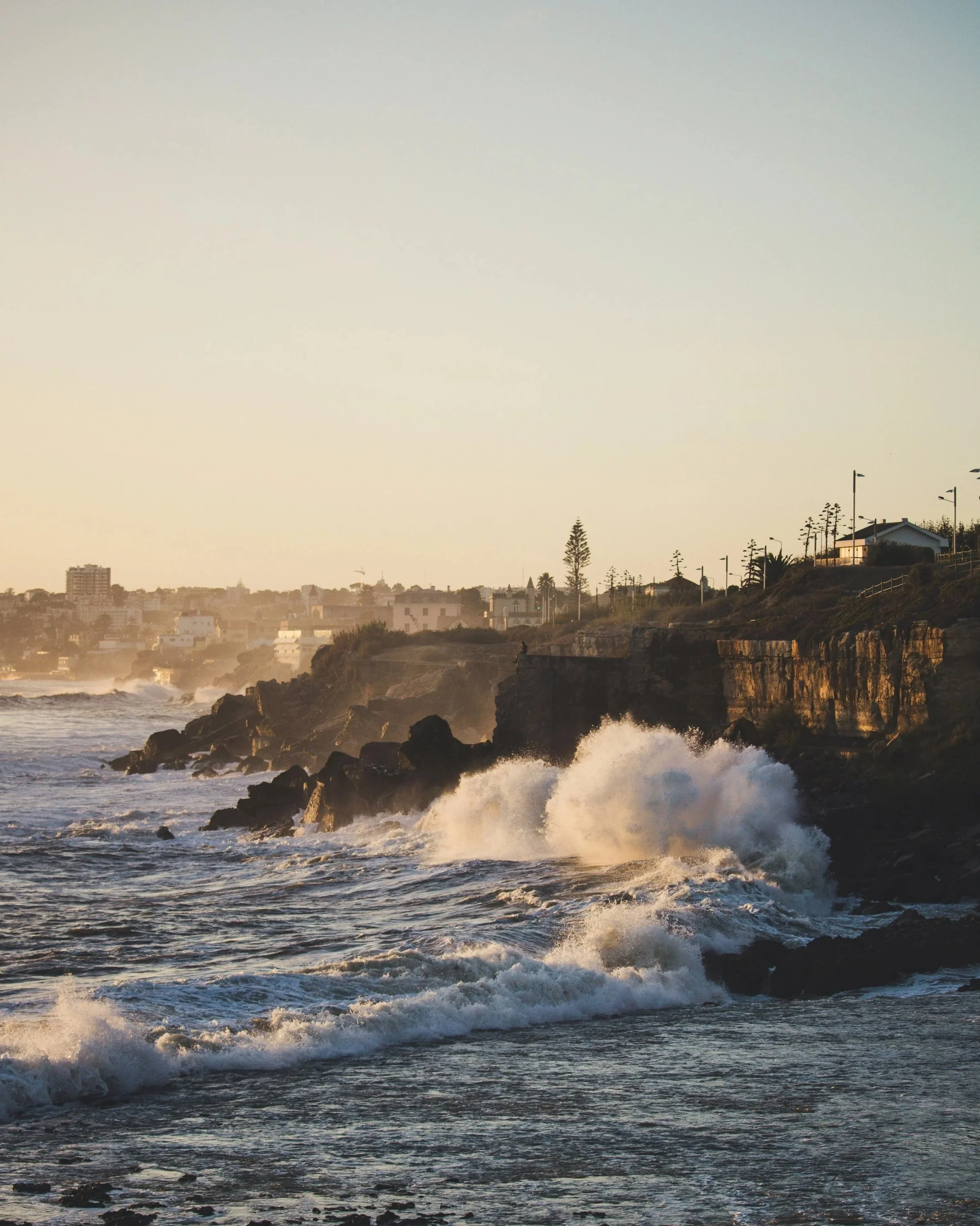 Waves crashing against rocky shoreline at sunset with houses and trees on a cliff in the background.
