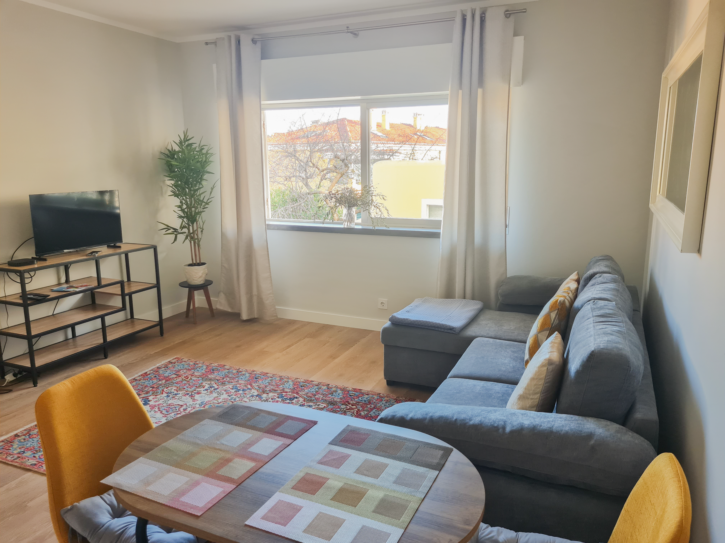 Living room with a gray sectional sofa, a round wooden dining table with colorful placemats and yellow chairs, a black TV on a wooden stand, a potted plant, beige curtains, and a window with an outside view of rooftops and trees.