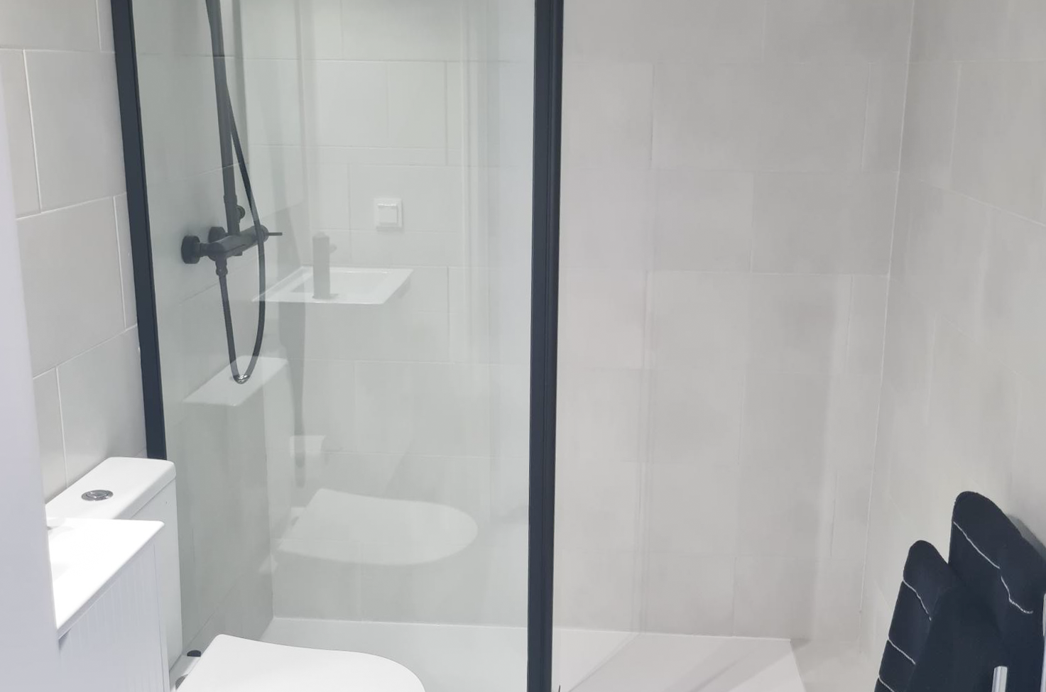 Modern bathroom with walk-in shower enclosed by glass, black shower fixtures, a small white sink, and a black towel hanging on a rack.