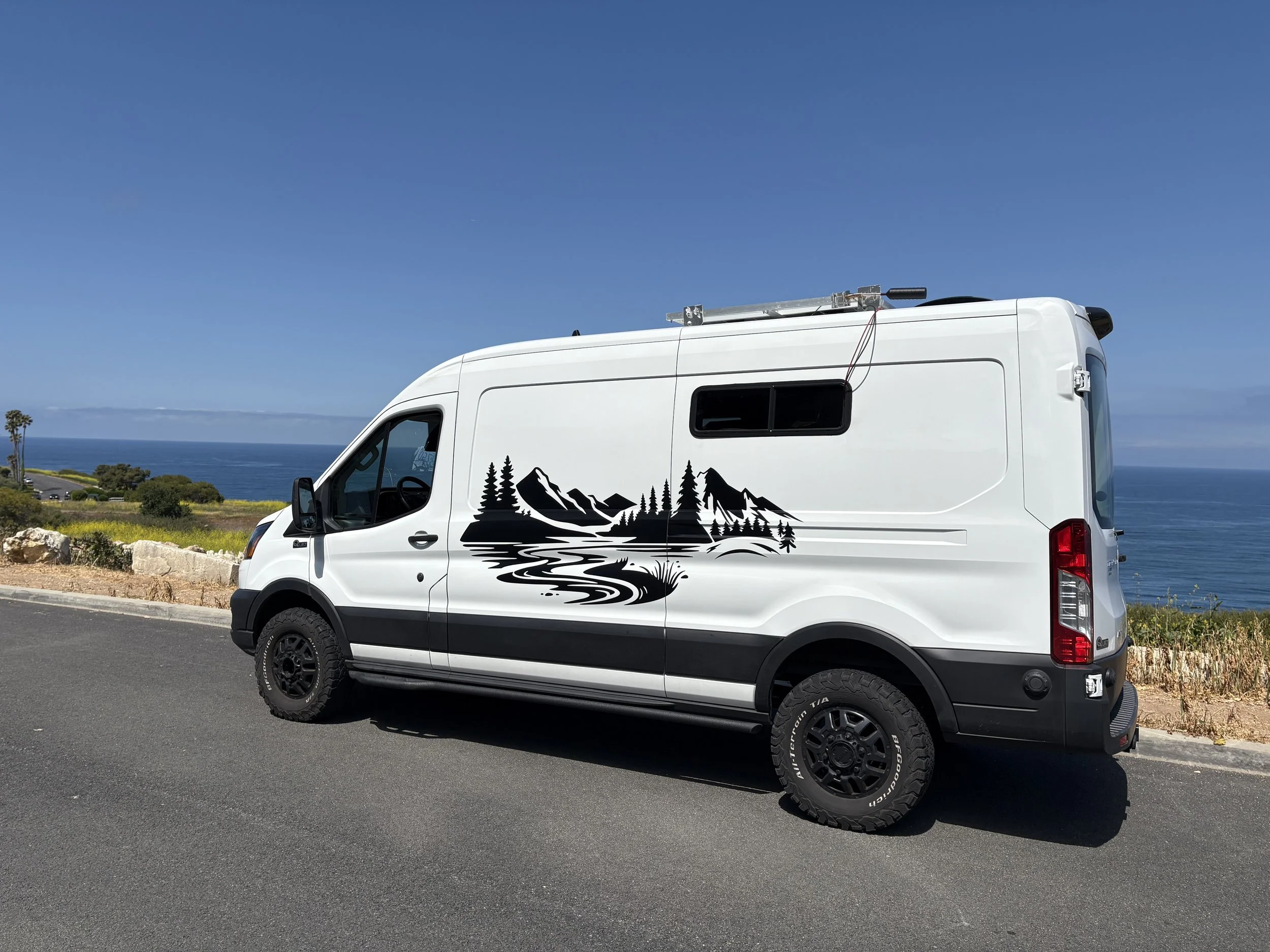 White camper van with mountain and forest decal parked by the roadside near the coast