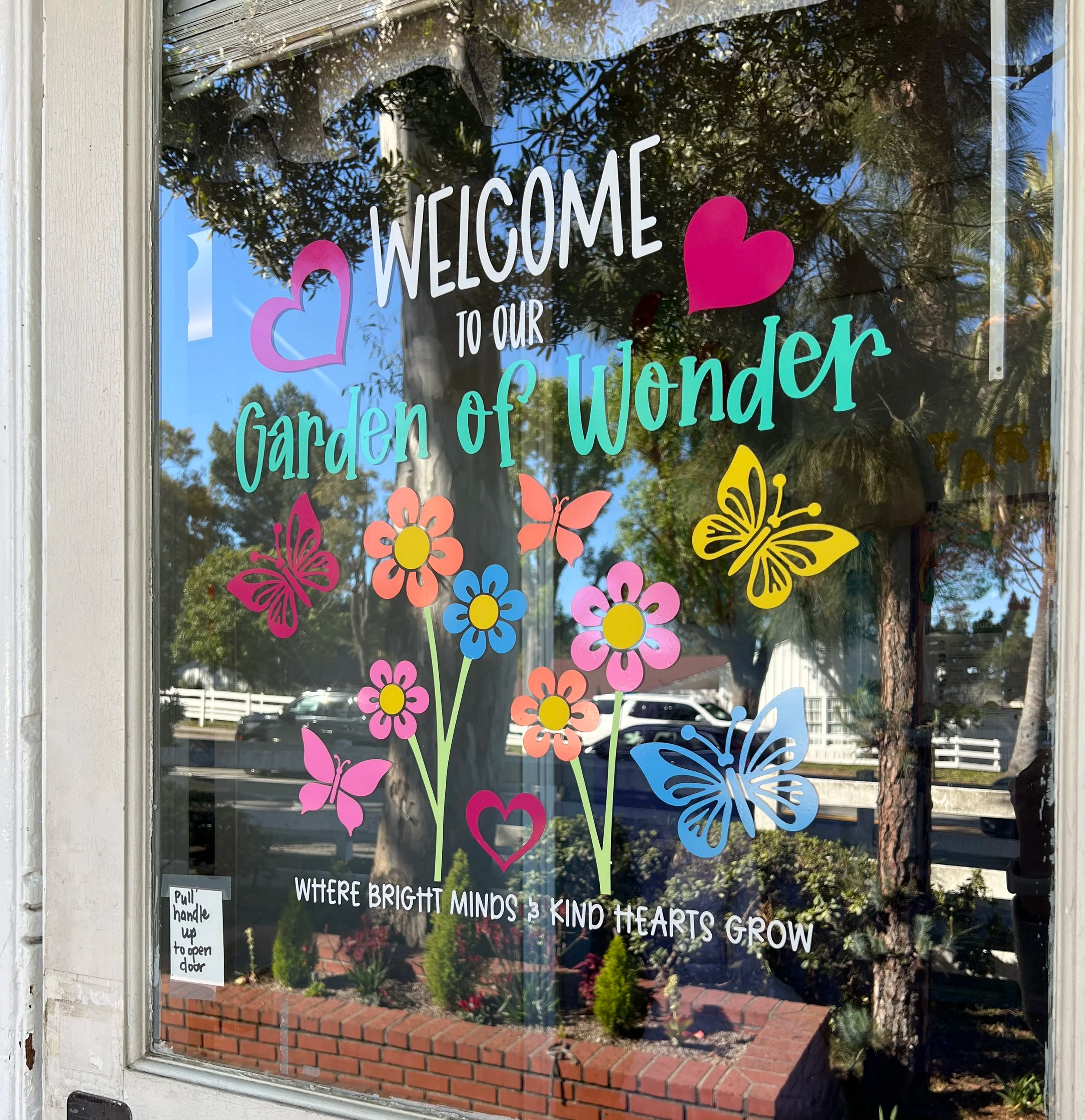 Decorative window sign welcoming visitors to the Garden of Wonder. The sign features colorful flowers, butterflies, and hearts, with a cheerful quote about bright minds and kind hearts. The window reflects trees, sky, and parked cars outside.
