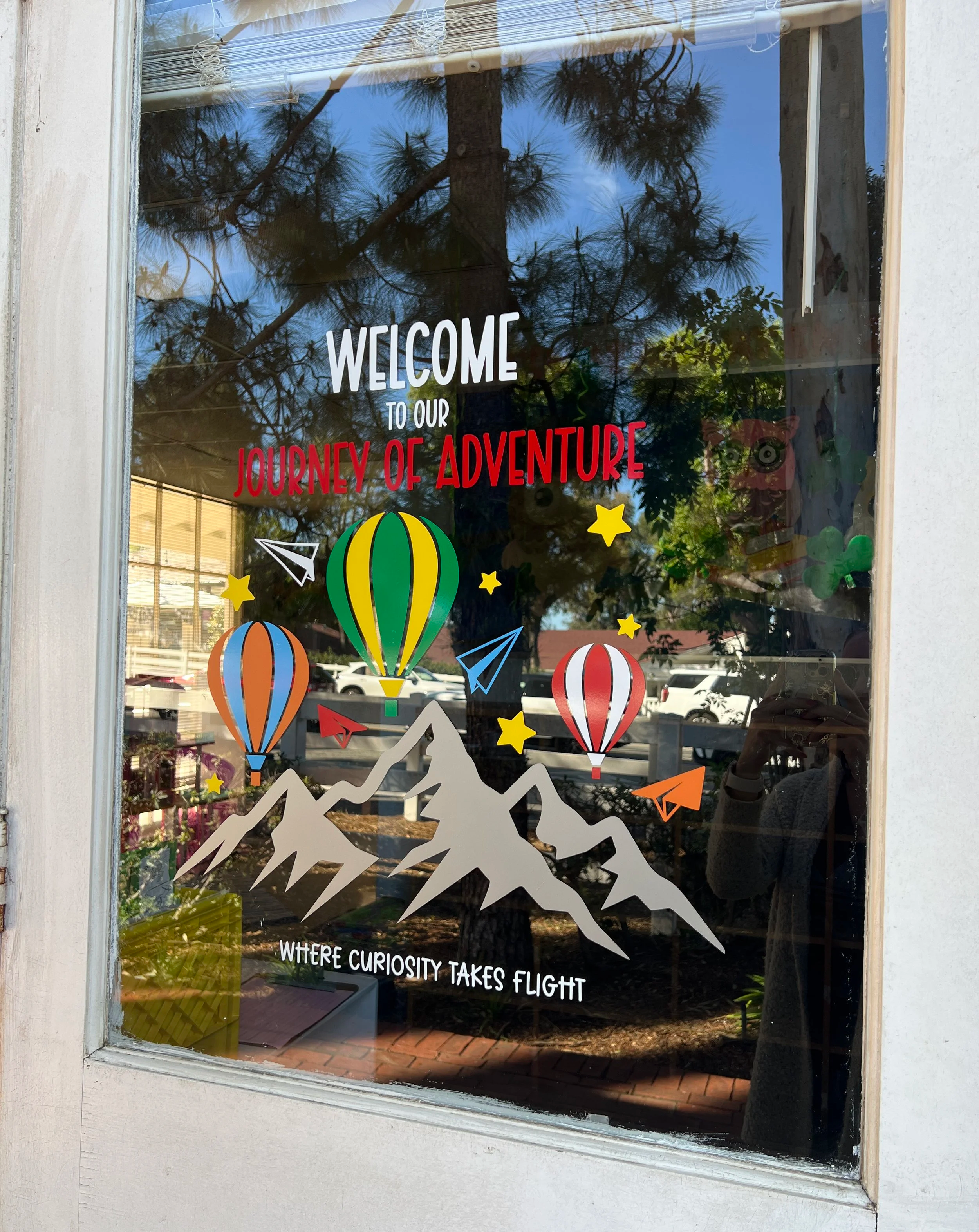 Decorative window poster featuring mountains, hot air balloons, paper airplanes, stars, and the text 'Welcome to our Journey of Adventure' and 'Where curiosity takes flight'.