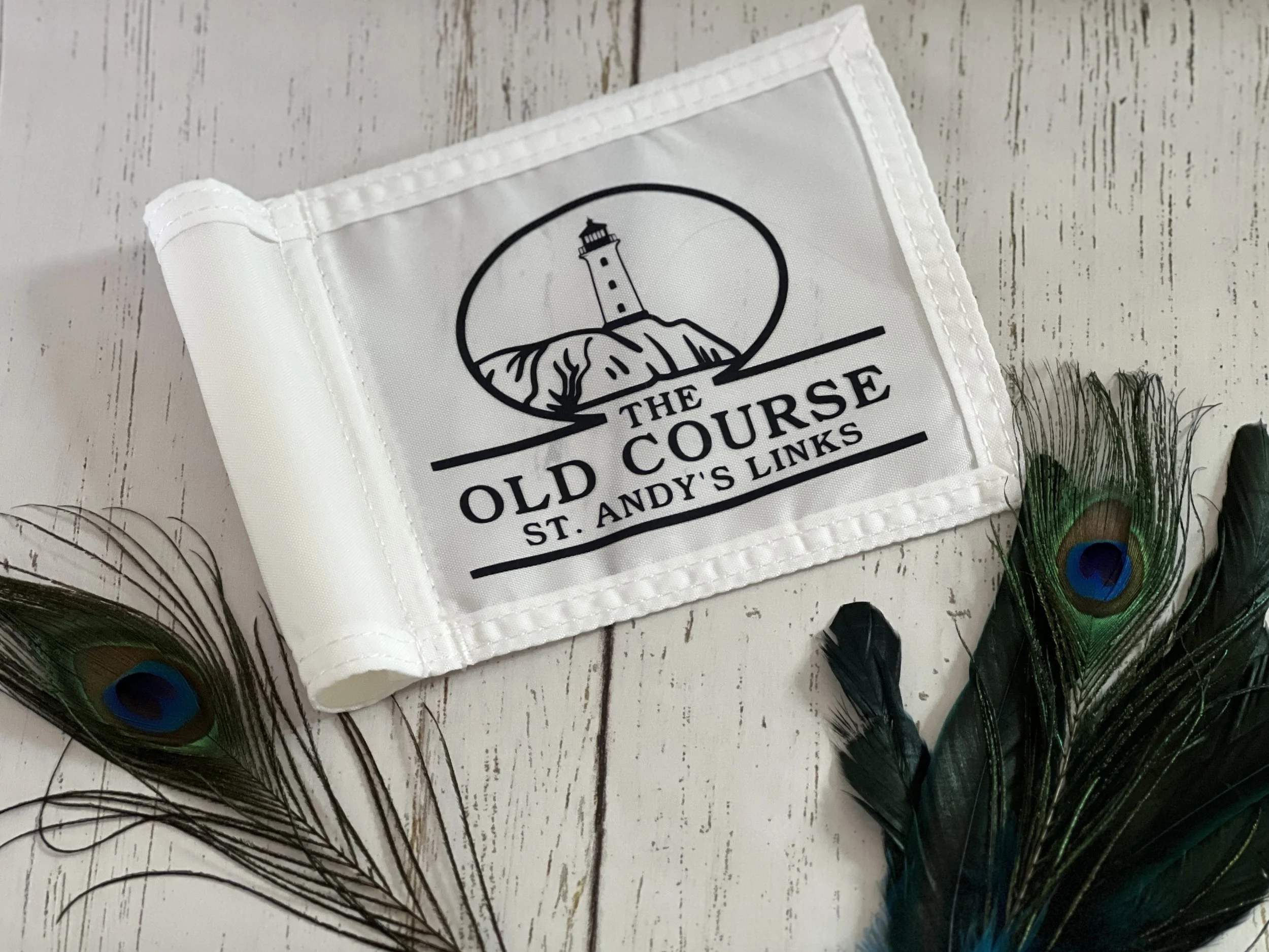 White flag with black logo and text, placed on a white wooden surface, surrounded by peacock feathers.