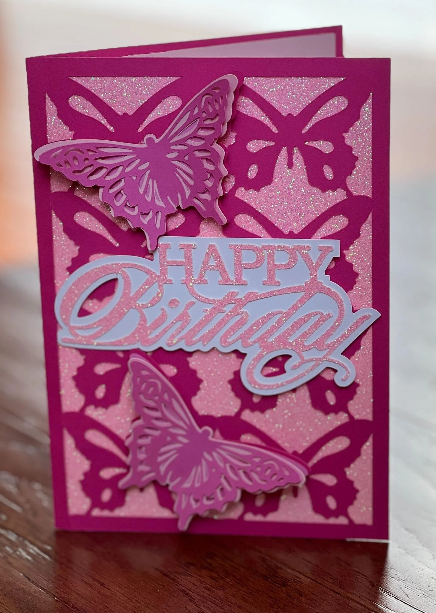 Pink birthday card with butterfly cutouts, glitter background, and a white and pink "Happy Birthday" message.