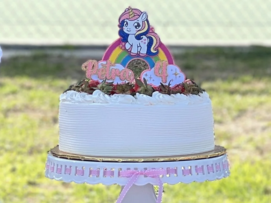 A birthday cake decorated with a rainbow and a My Little Pony character on top, with pink lettering spelling "Petra 4" and small decorative clouds, set against a natural outdoor background.