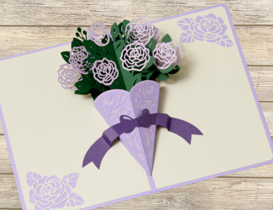A greeting card with a 3D paper bouquet of violet and lavender flowers and green leaves, tied with a purple ribbon, on a light wood surface.