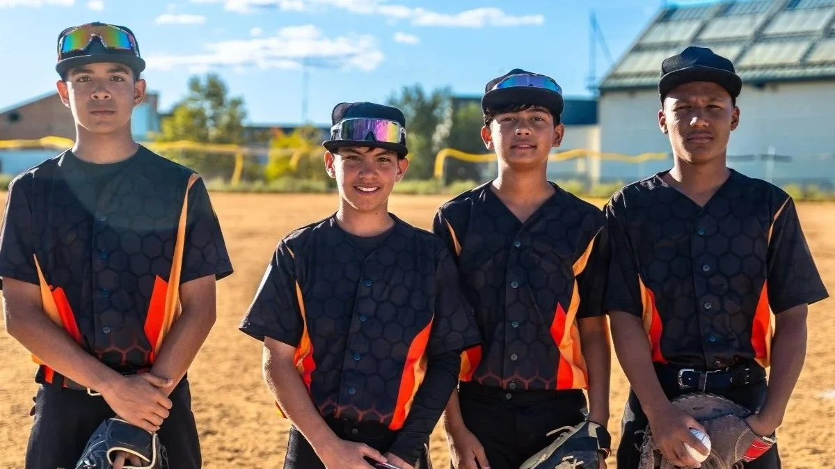 Ultimate Goal Book Four young boys in black and orange baseball uniforms standing on a baseball field during daytime, some holding baseball gloves and ball caps.