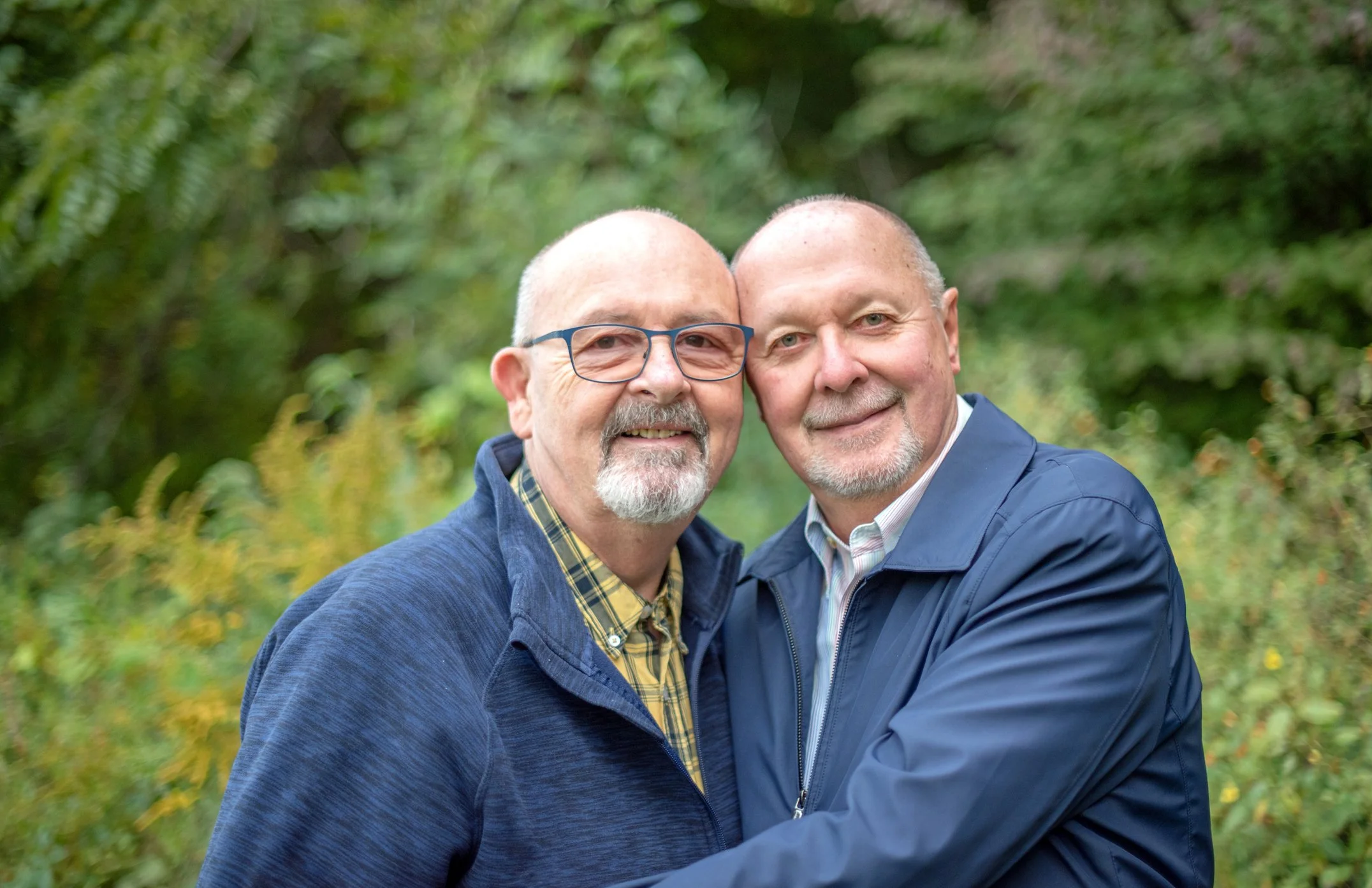 Two middle-aged men hugging outdoors, smiling, with a background of green foliage.