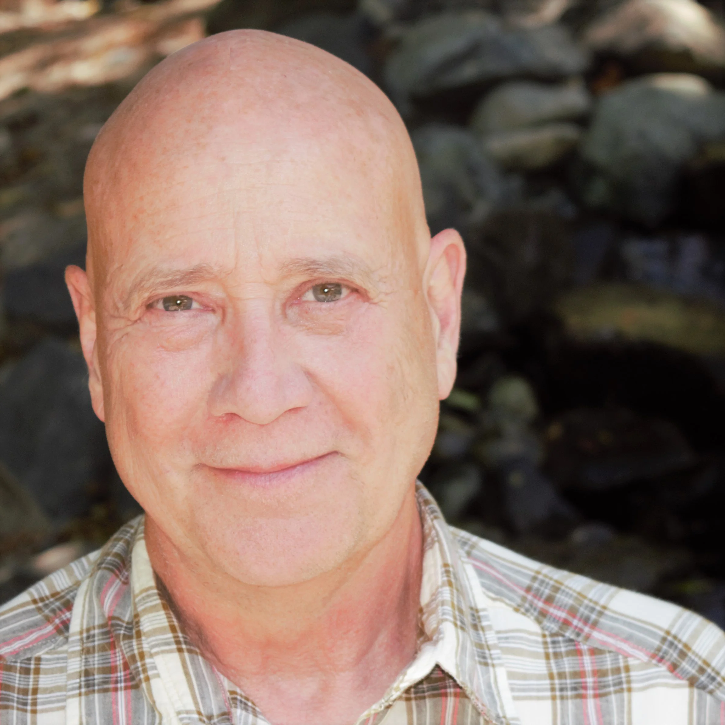 Close-up photo of smiling middle-aged man with a bald head, wearing a plaid shirt, outdoors with rocks in the background.