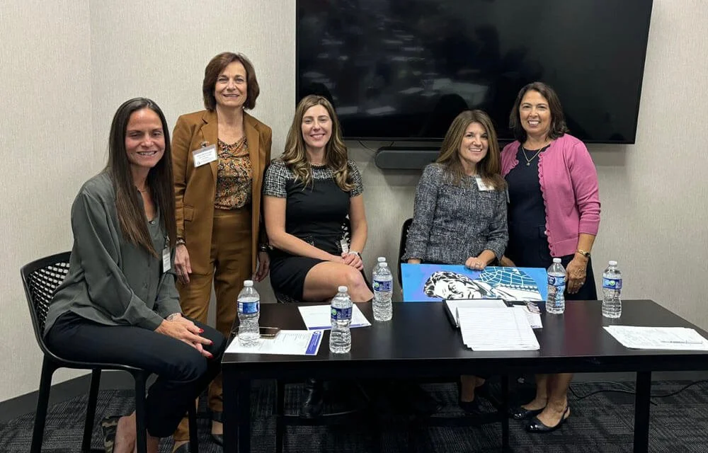 Five women in business attire, sitting and standing around a conference table in a meeting room.