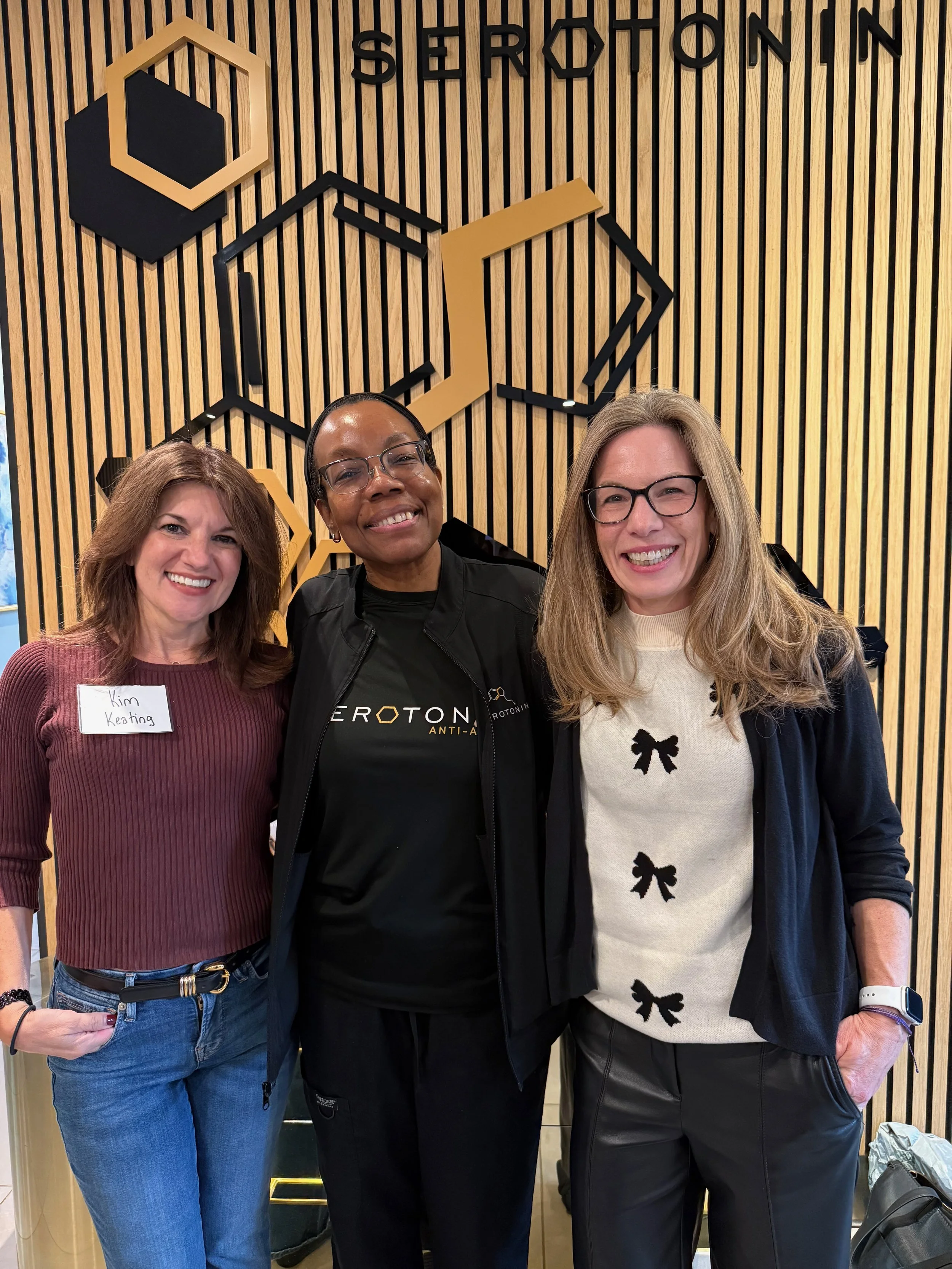 Three women standing together indoors in front of a wooden wall with black and yellow geometric decorations and the word 'SORTON' displayed on the wall. The woman on the left has brown hair, wears a brown ribbed top, blue jeans, and a name tag that reads 'Kim Keating.' The woman in the middle has dark skin, glasses, a black jacket, and a black shirt. The woman on the right has blonde hair, glasses, and is wearing a beige sweater with black bow designs and a black jacket.