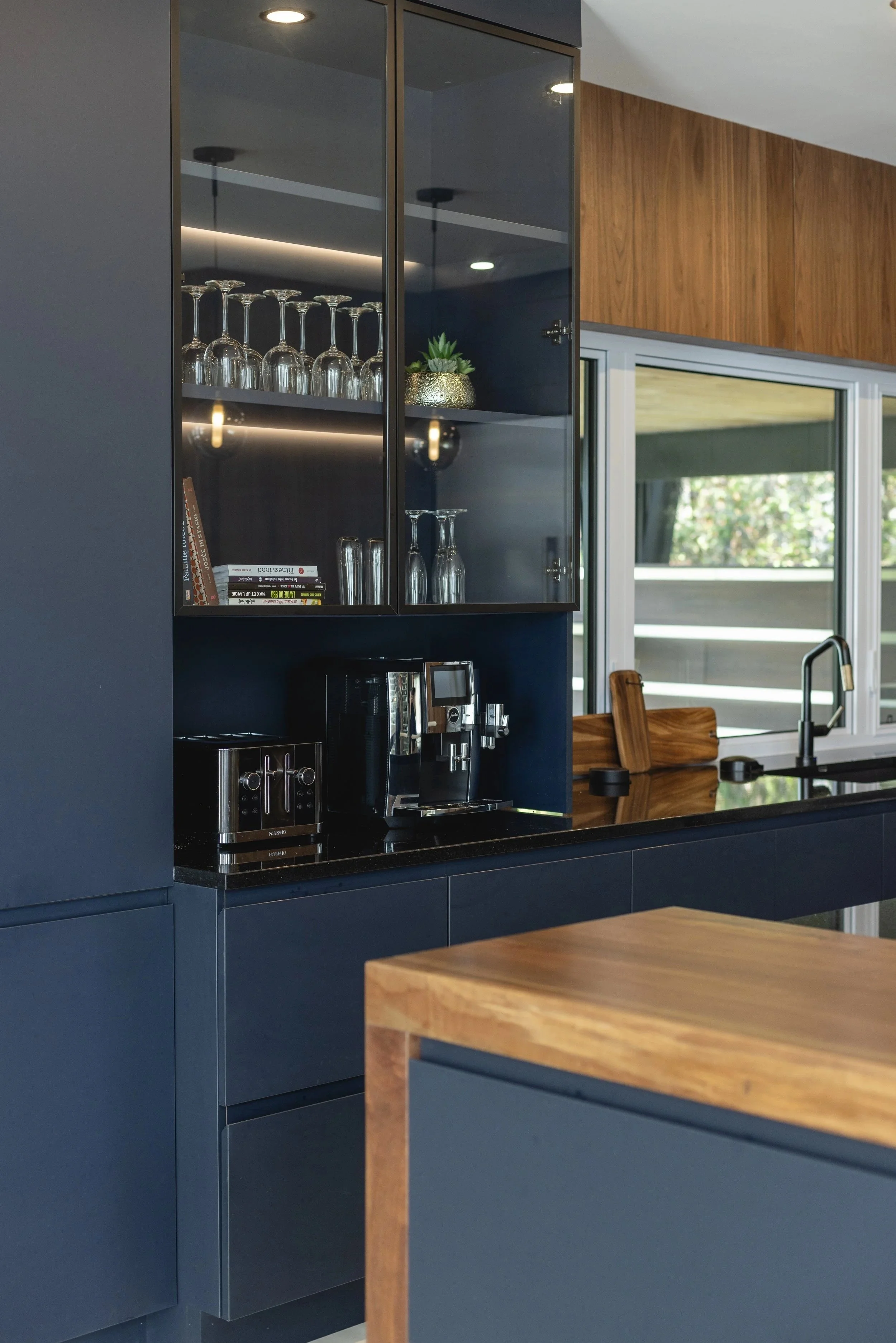 Modern kitchen with navy blue cabinets, a black countertop, and wooden accents. Glassware and books are displayed in a glass-front cabinet. Coffee machine, toaster, and kitchen sink visible near window with outdoor view.