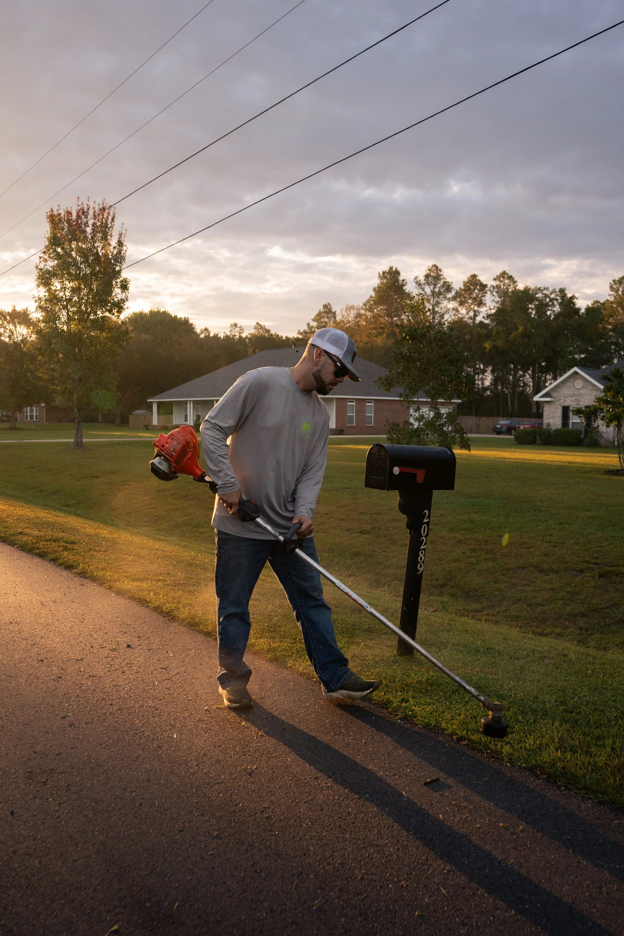 Mike Genesse of Genesse Lawn & Landscaping using a string trimmer along the curb of a residential street during sunset.