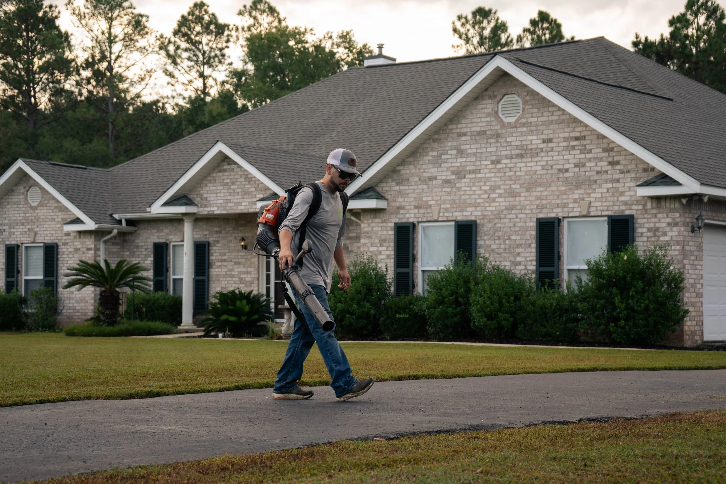 Mike Genesse of Genesse Lawn & Landscaping walking on sidewalk near a brick house, carrying outdoor equipment, wearing sunglasses, a cap, and backpack.