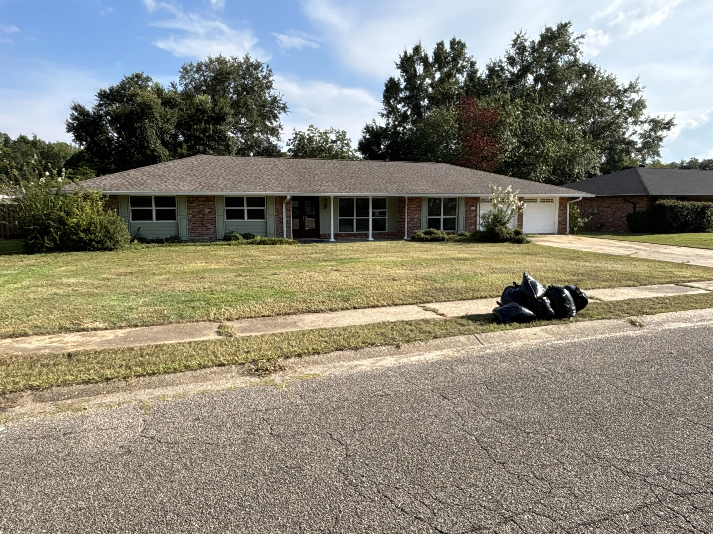 A single-story ranch house with a brick and light siding exterior, front lawn, and a driveway with garbage bags in front, against a backdrop of trees and a blue sky with some clouds.