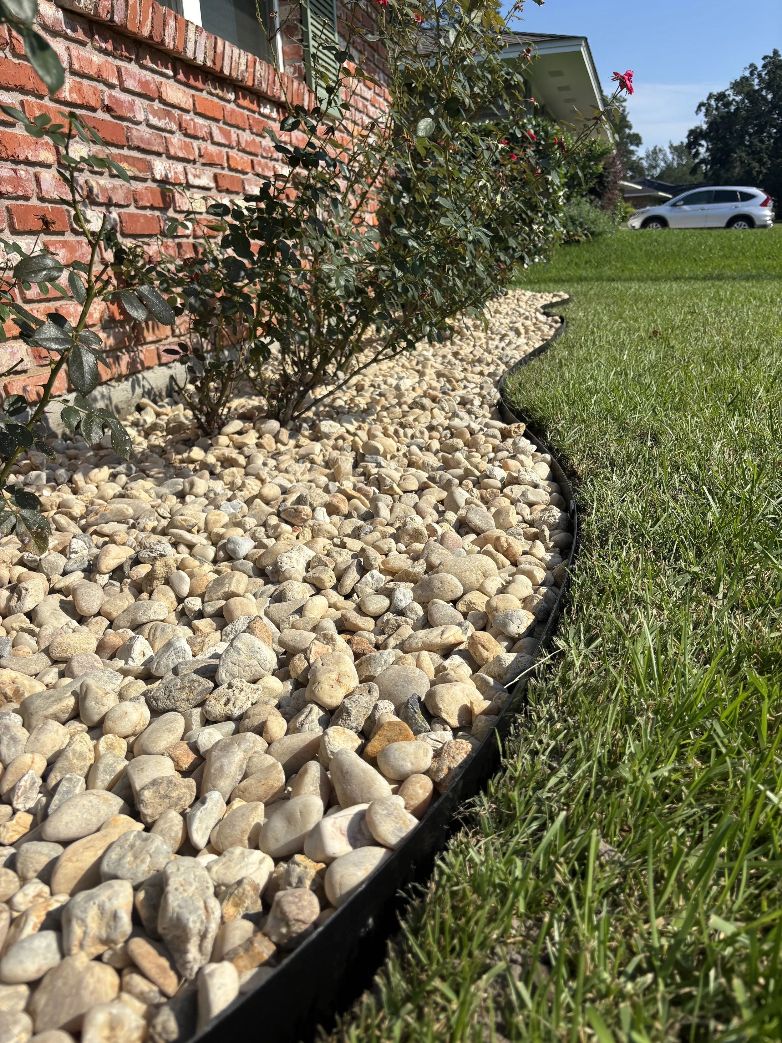 A landscaped yard with a brick house wall on the left, a row of small green bushes, a gravel bed separated by a black edging, and a grassy lawn on the right. A silver car is parked in the driveway in the background.
