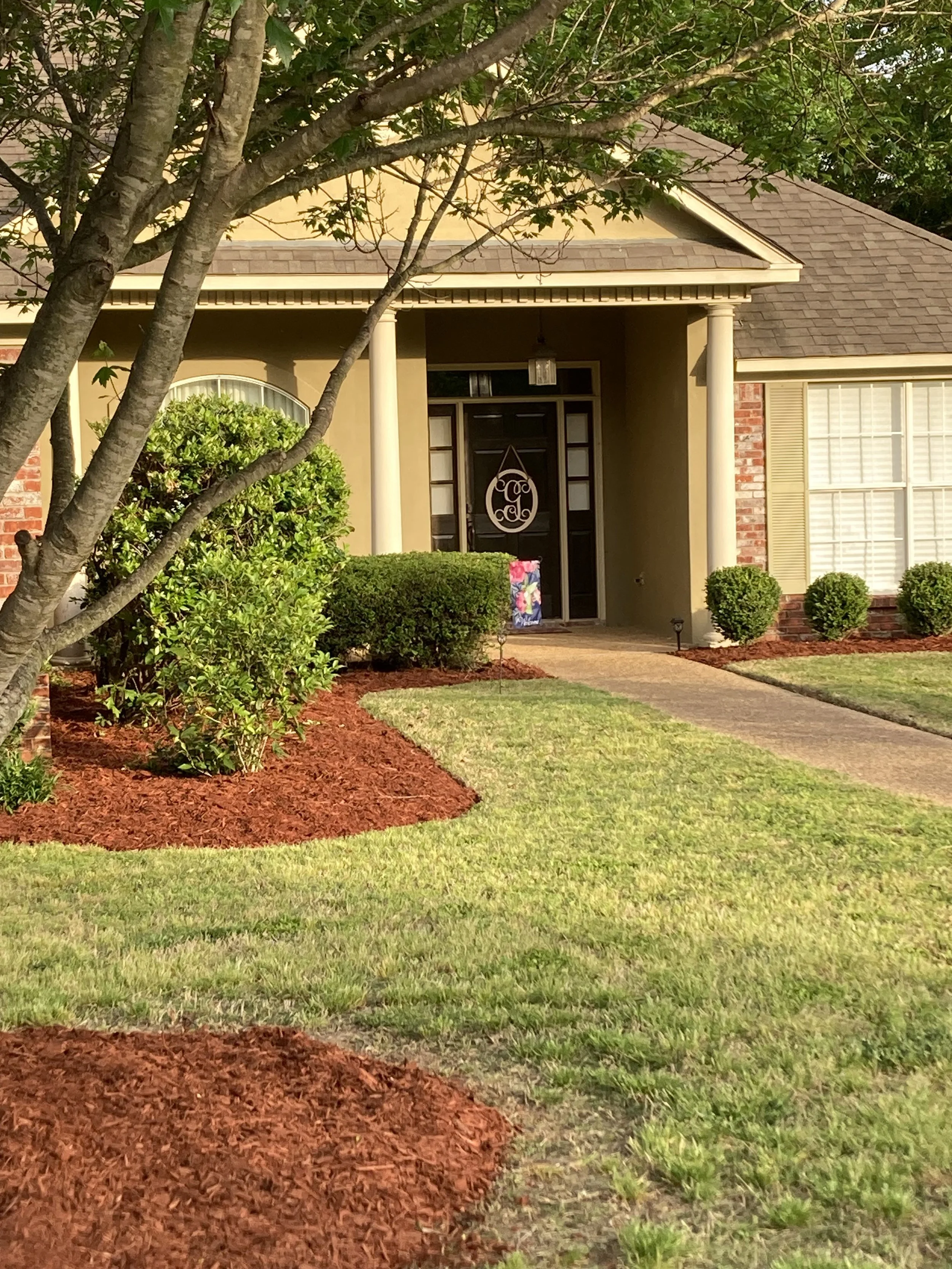 Front yard of a house with a pathway leading to the front door, surrounded by neatly trimmed bushes and a tree in the foreground. The house has a covered porch with two white columns, a black door with decorative glass, and a window with blinds on th