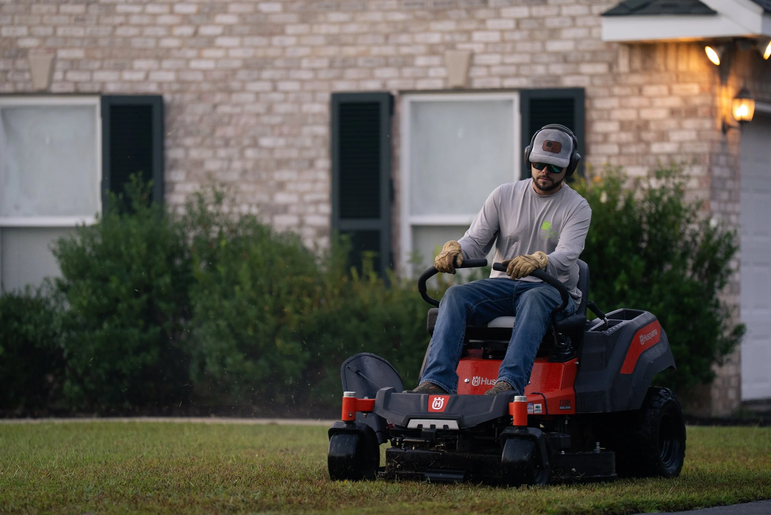 Mike Genesse of Genesse Lawn & Landscaping operating a riding lawn mower on a lawn in front of a brick house with windows and bushes.
