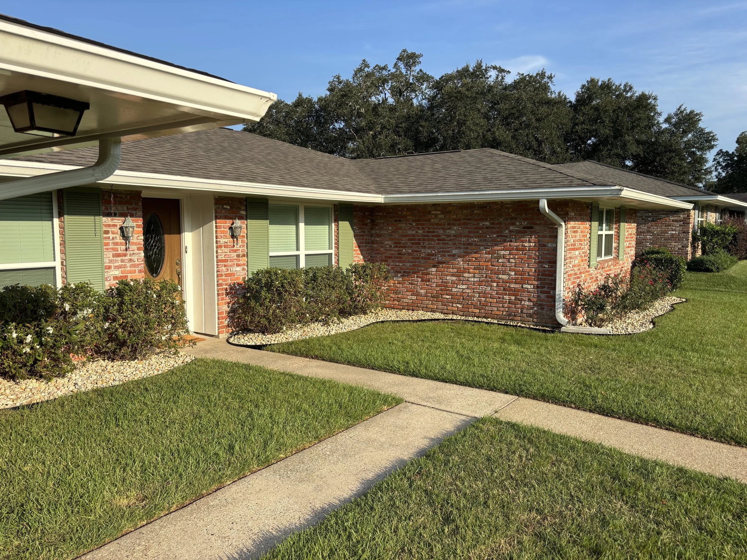 A brick house with green shutters and a gray roof, surrounded by a green lawn, bushes, and a walkway leading to the front door, under a blue sky.
