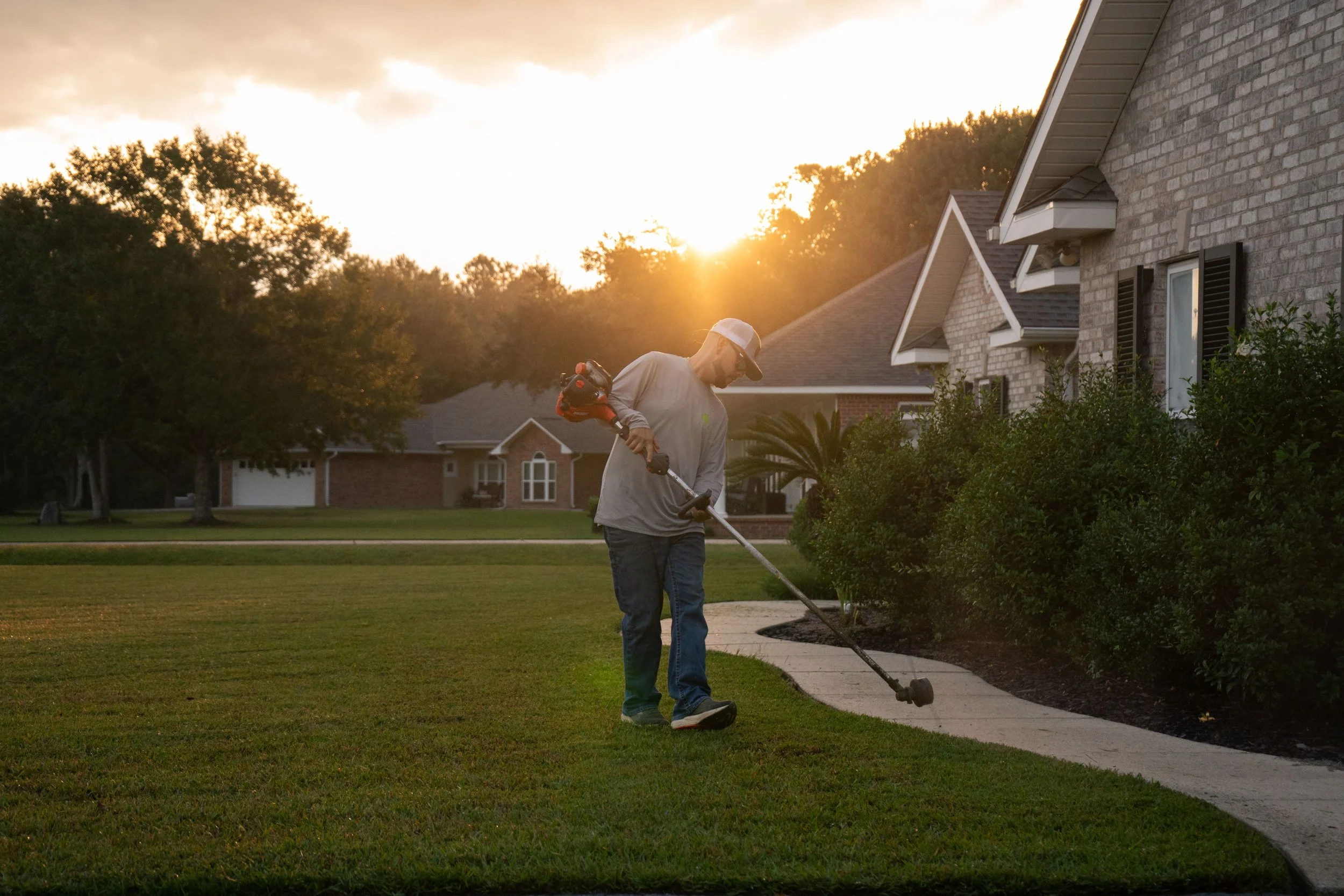 Mike Genesse of Genesse Lawn & Landscaping trimming bushes with a weed trimmer on the front lawn of a suburban house at sunset.