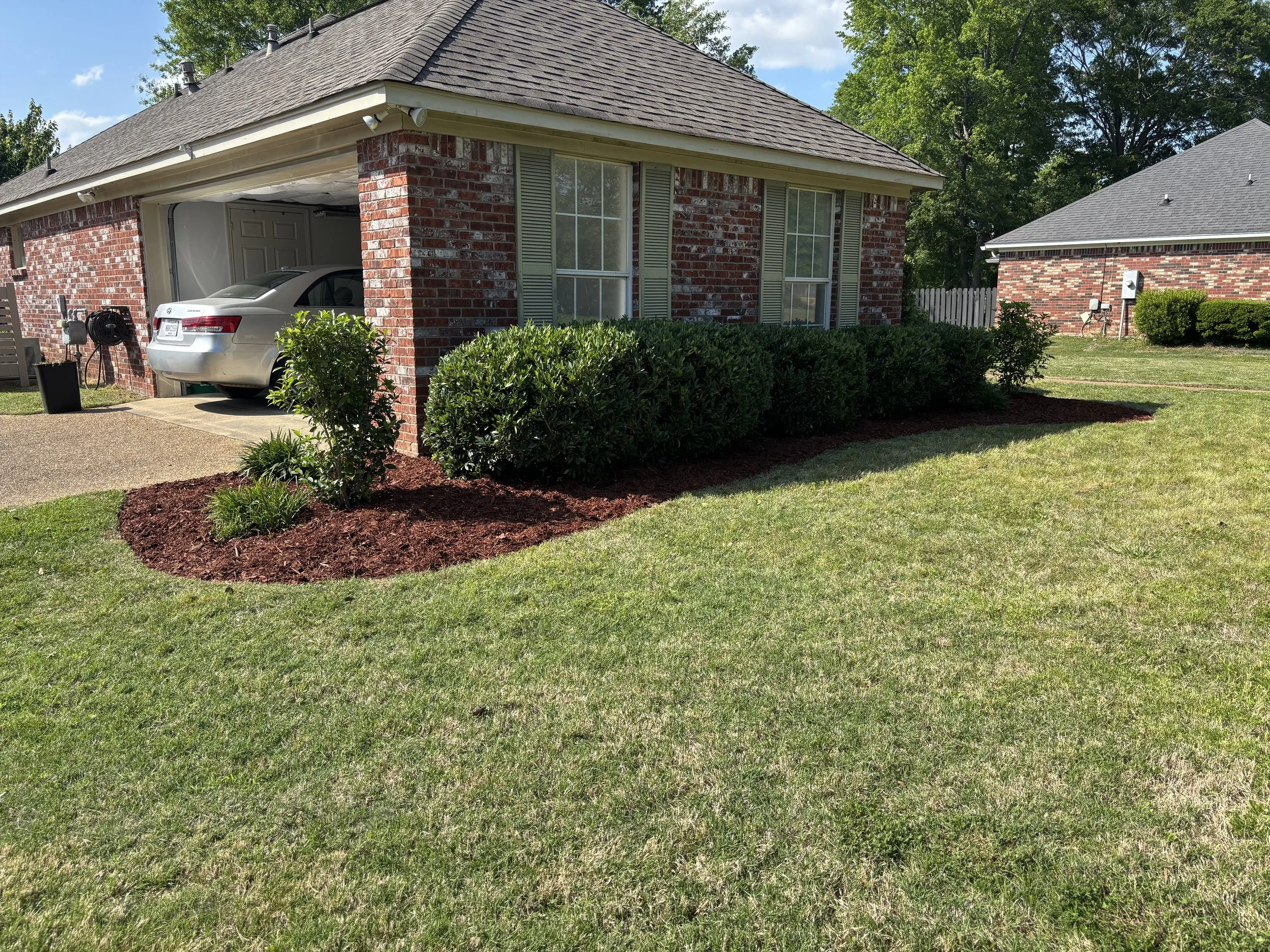 A suburban house with a brick exterior and green shutters. A landscaped garden bed with bushes and mulch is in front of the house. A silver car is parked in the garage, which is open. The lawn is green and well-maintained, and there are trees in the