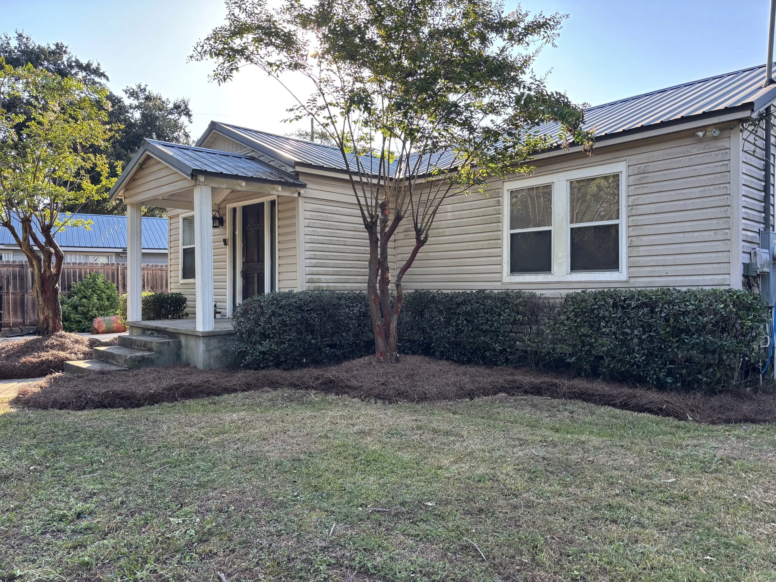 Front yard of a house with light-colored siding, small porch with stairs, black door, and two windows. Landscaping includes trees and bushes along the house, with freshly mulched flower beds and a grass lawn in Long Beach, MS.