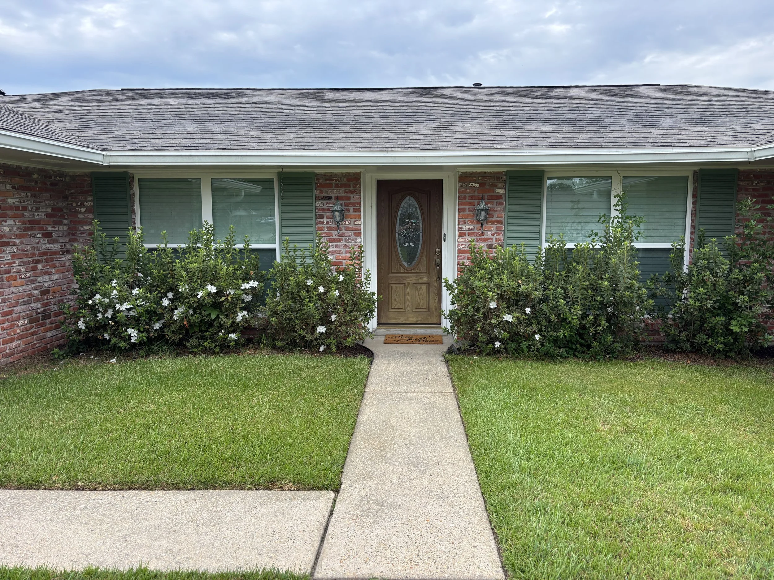 Front view of a brick house with a brown door, green shutters, two lanterns beside the door, and bushes with white flowers in front of the house. A concrete sidewalk leads to the front door and a well-maintained lawn surrounds it.