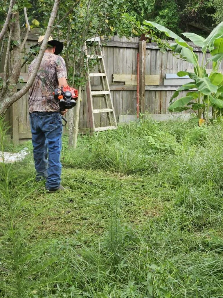 Mike Genesse of Genesse Lawn & Landscaping in camouflage shirt and jeans with a leaf blower in a backyard surrounded by green grass, trees, and a wooden fence, with a ladder leaning against the fence.
