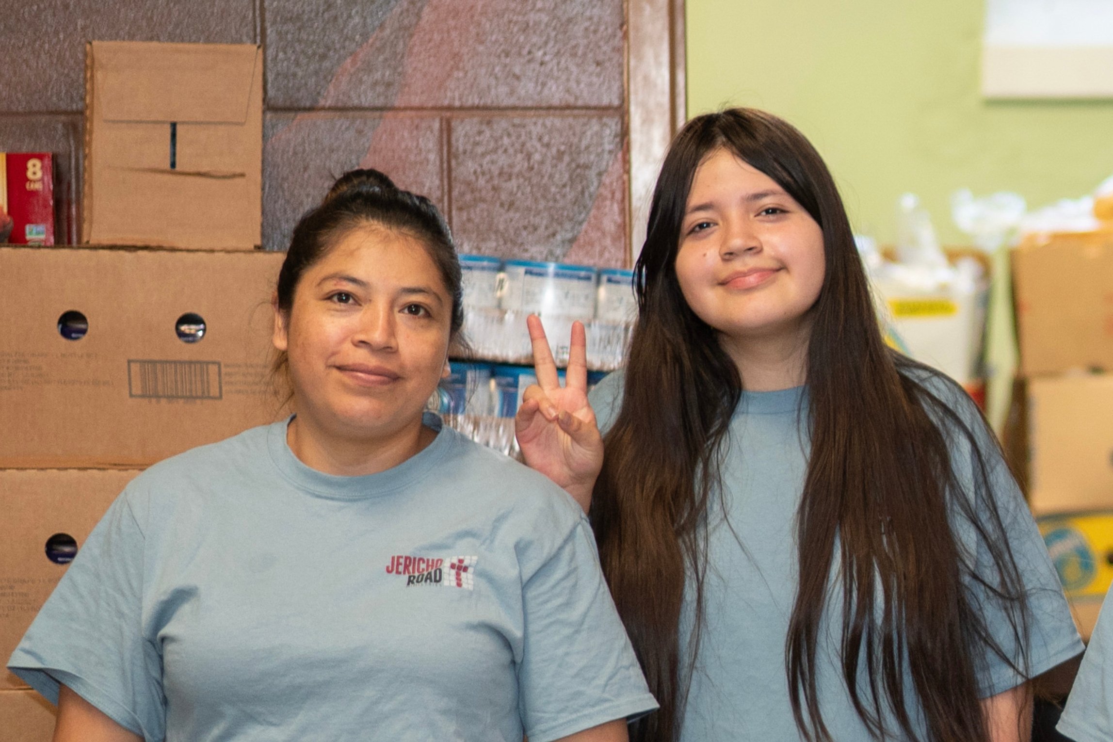 Two women in light blue shirts standing in front of stacked cardboard boxes at a food pantry, smiling, with one girl making a peace sign behind the other.