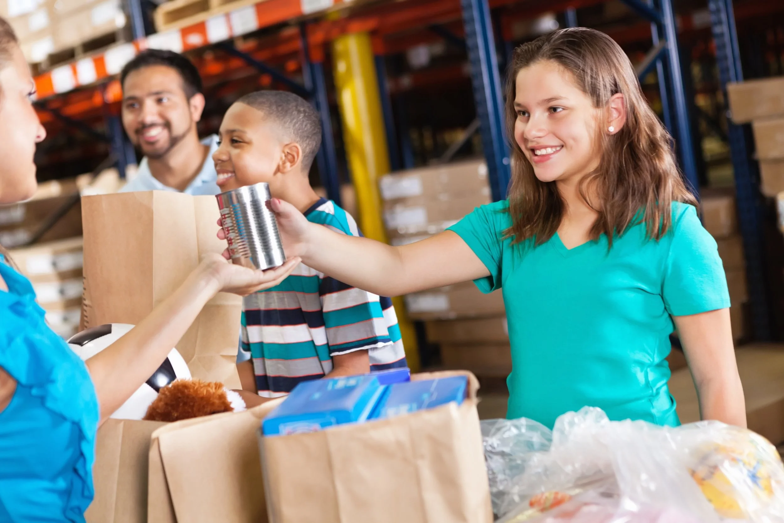 A girl in a turquoise shirt hands canned food to a woman at a food bank, with children and a man in the background.