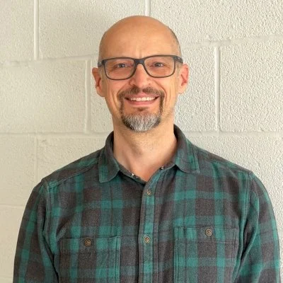 Photo of Justin Eccles, Jericho Road's Fundraising and Marketing Manager, wearing a plaid shirt standing in front of a white brick wall.