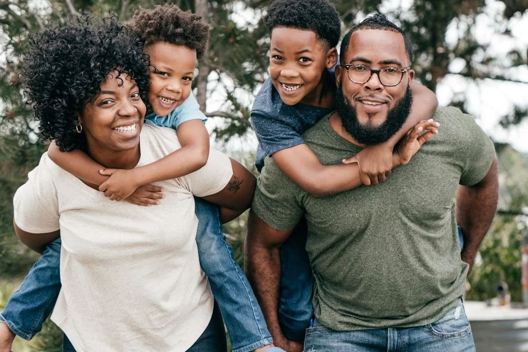 A happy family of four outdoors, with two adults and two children, smiling and enjoying a playful moment.
