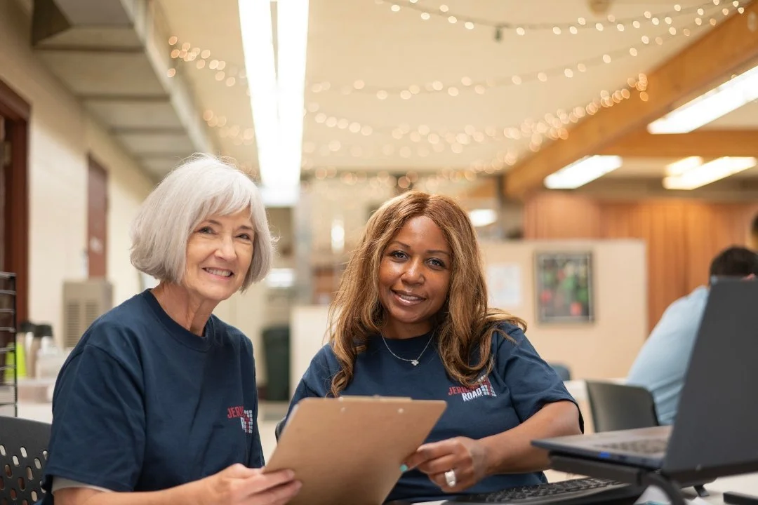 Two women sitting at a table, smiling at the camera. One holds a clipboard; they are in a room decorated with string lights at Jericho Road Ministries' Elliot Park location.