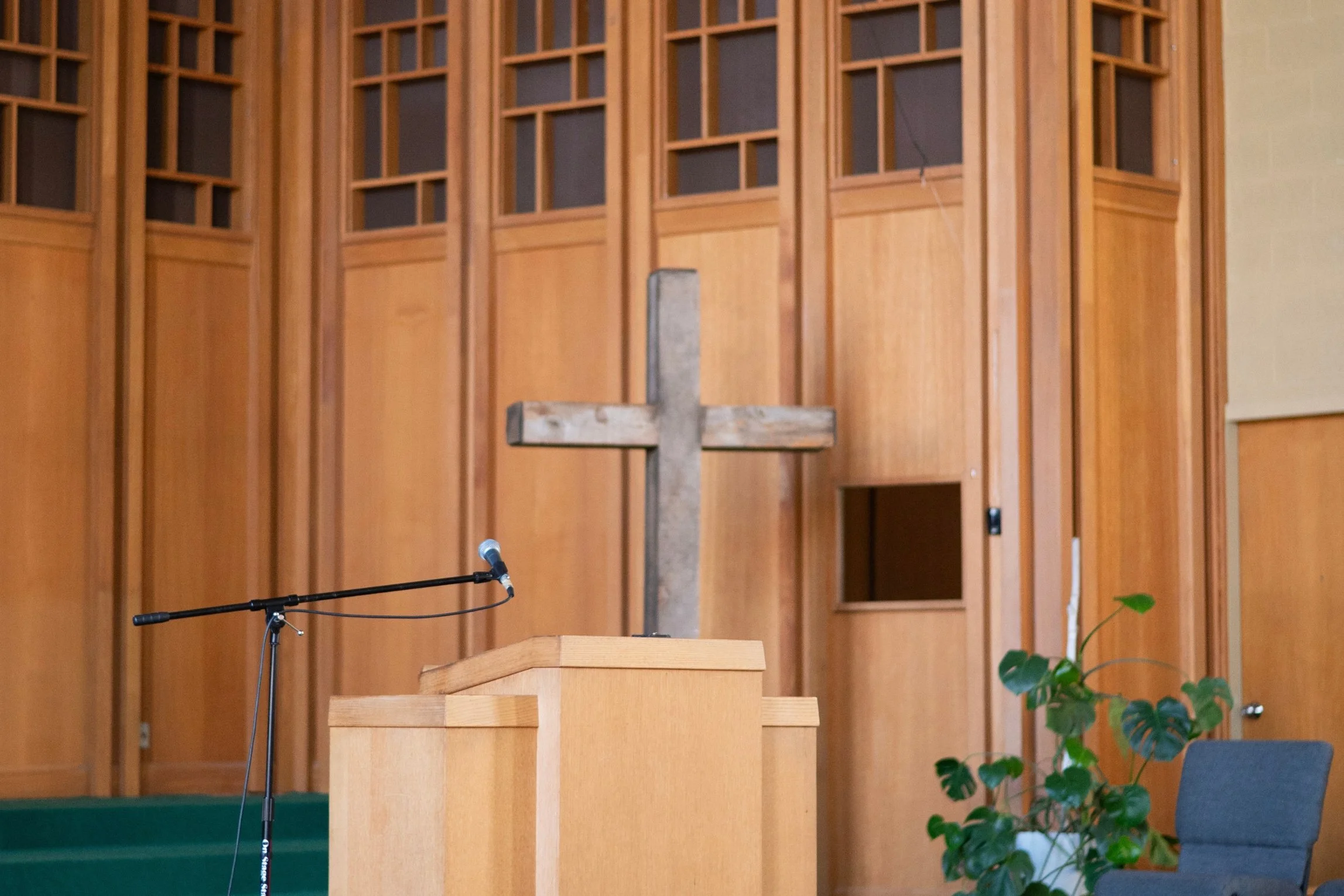 A wooden cross stands behind a podium inside a church, with a microphone on a stand nearby. The background features wooden panels and a potted plant with green leaves.