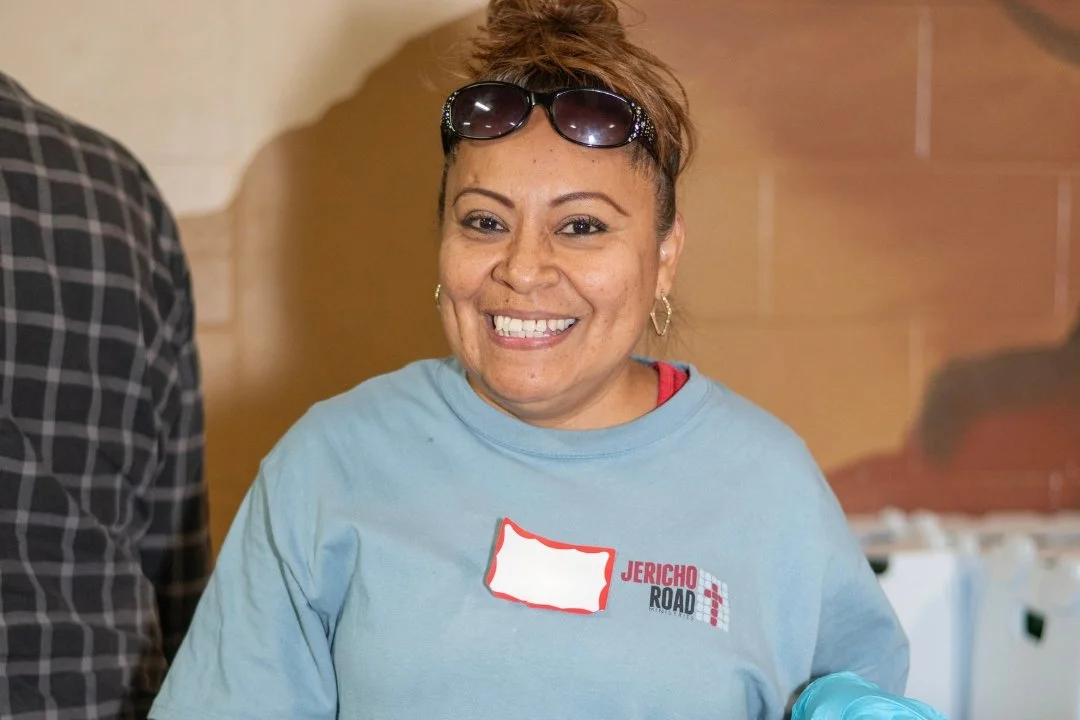 A smiling woman wearing a light blue Jericho Road t-shirt with sunglasses on her head, standing indoors against a beige wall at Jericho Road Ministries' Powderhorn location in Minneapolis, Minnesota.