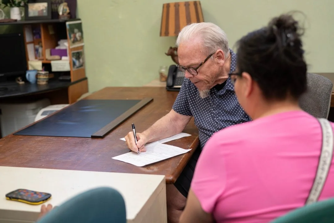 An elderly man who is a Jericho Road Ministries volunteer and a woman with her back to the camera at a wooden table, with the man filling out paperwork and the woman looking on.