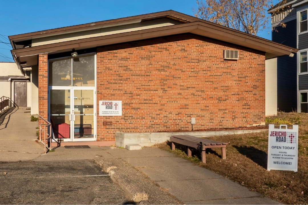 A small brick building with a glass entrance door, a sidewalk, and a bench outside. There are signs indicating the building is Jericho Road, open Tuesdays and Thursdays from 9:30 AM to 12:30 PM.