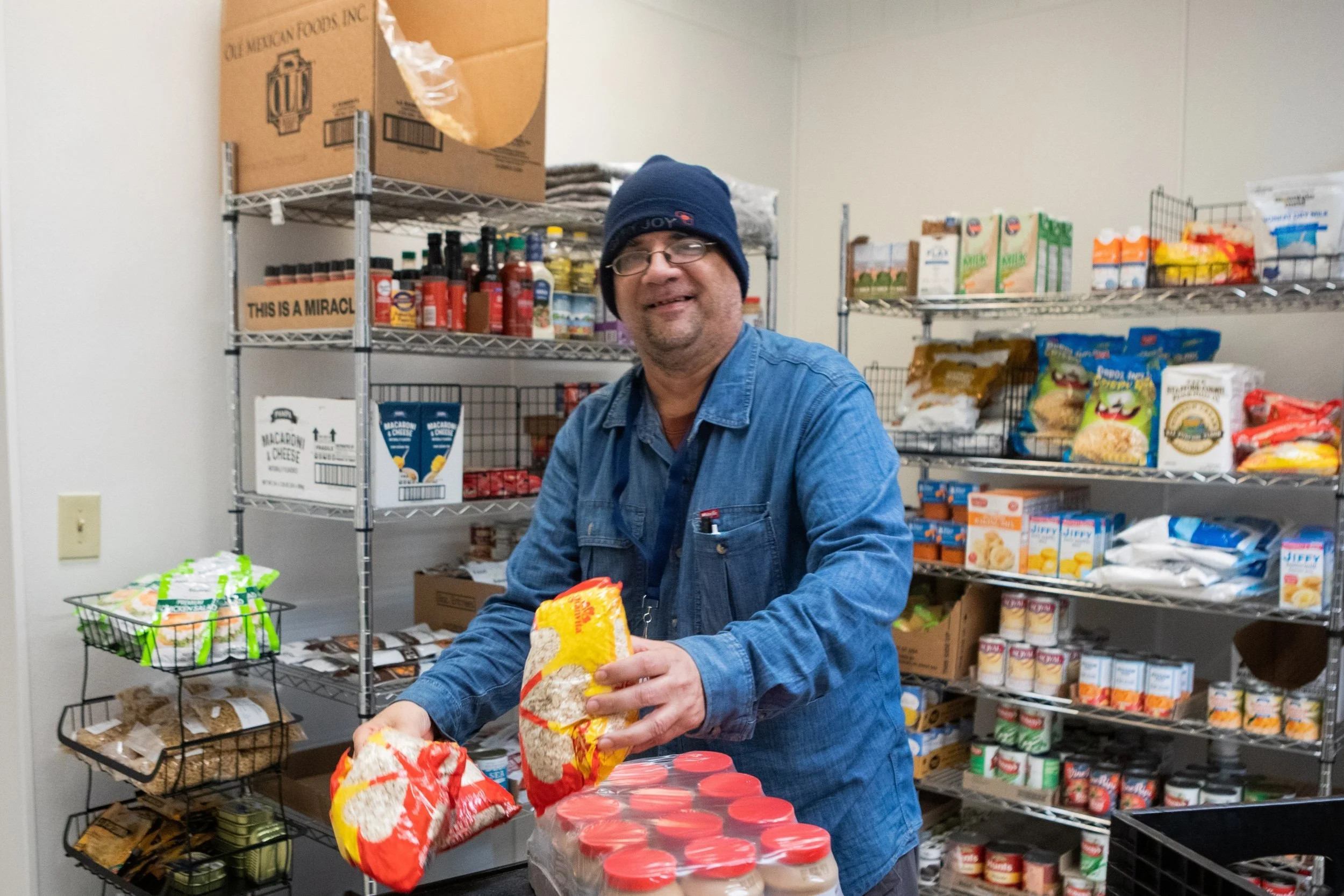 A man in a blue denim jacket smiling and holding a bag of non-perishable food, inside the food shelf at Jericho Road Ministries' Elliot Park location in Minneapolis with shelves of canned goods and other food supplies behind him.