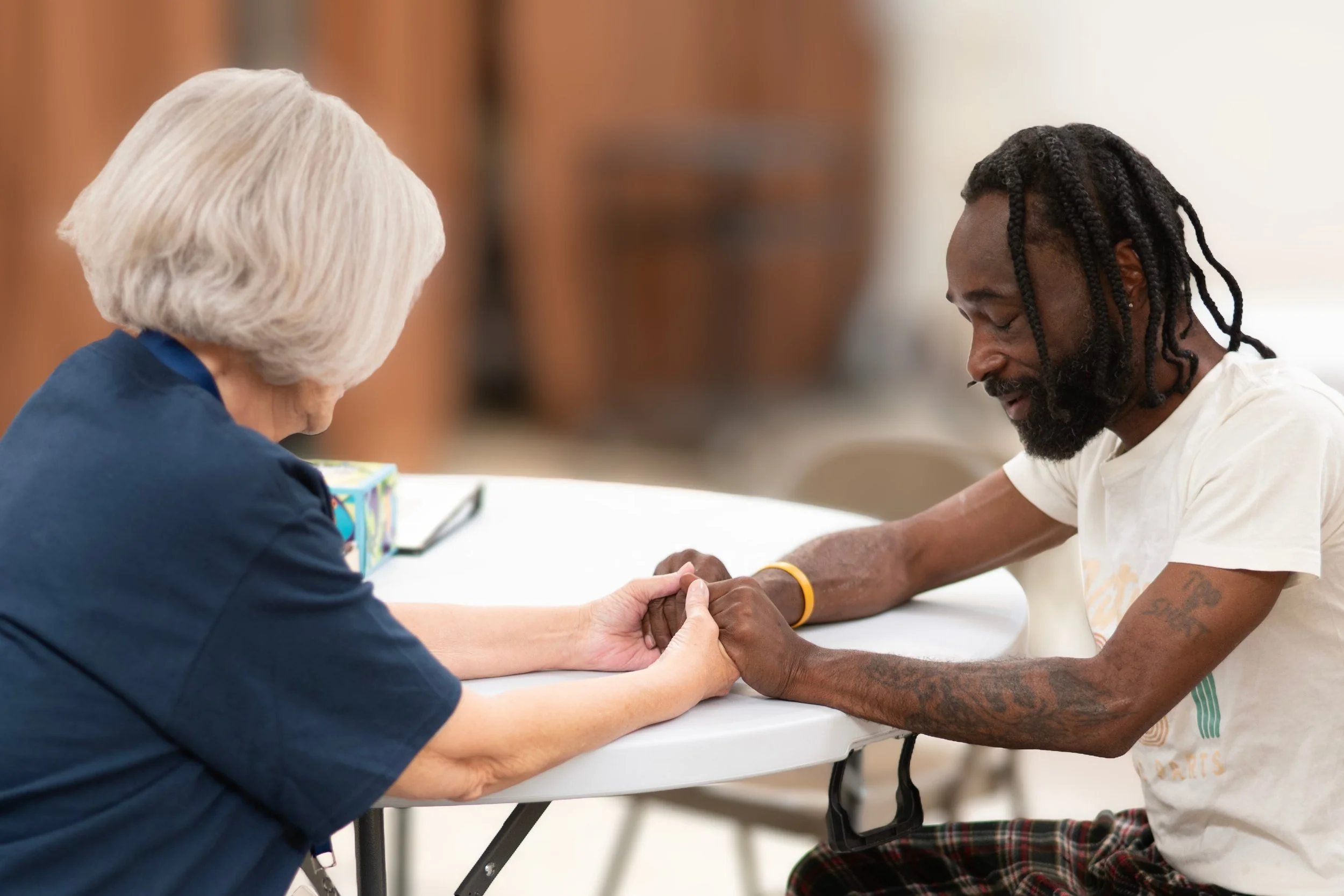 An elderly woman and a younger man praying together and holding hands at a table, sharing an emotional moment at Jericho Road Ministries in Minneapolis, Minnesota.