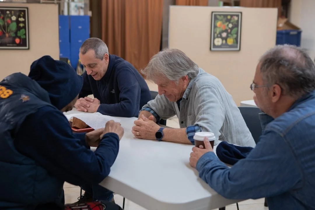 Four men sitting around a table, and praying together. The setting is indoors at Jericho Road Ministries' Elliot Park food pantry, with artwork on the walls and a neutral background.