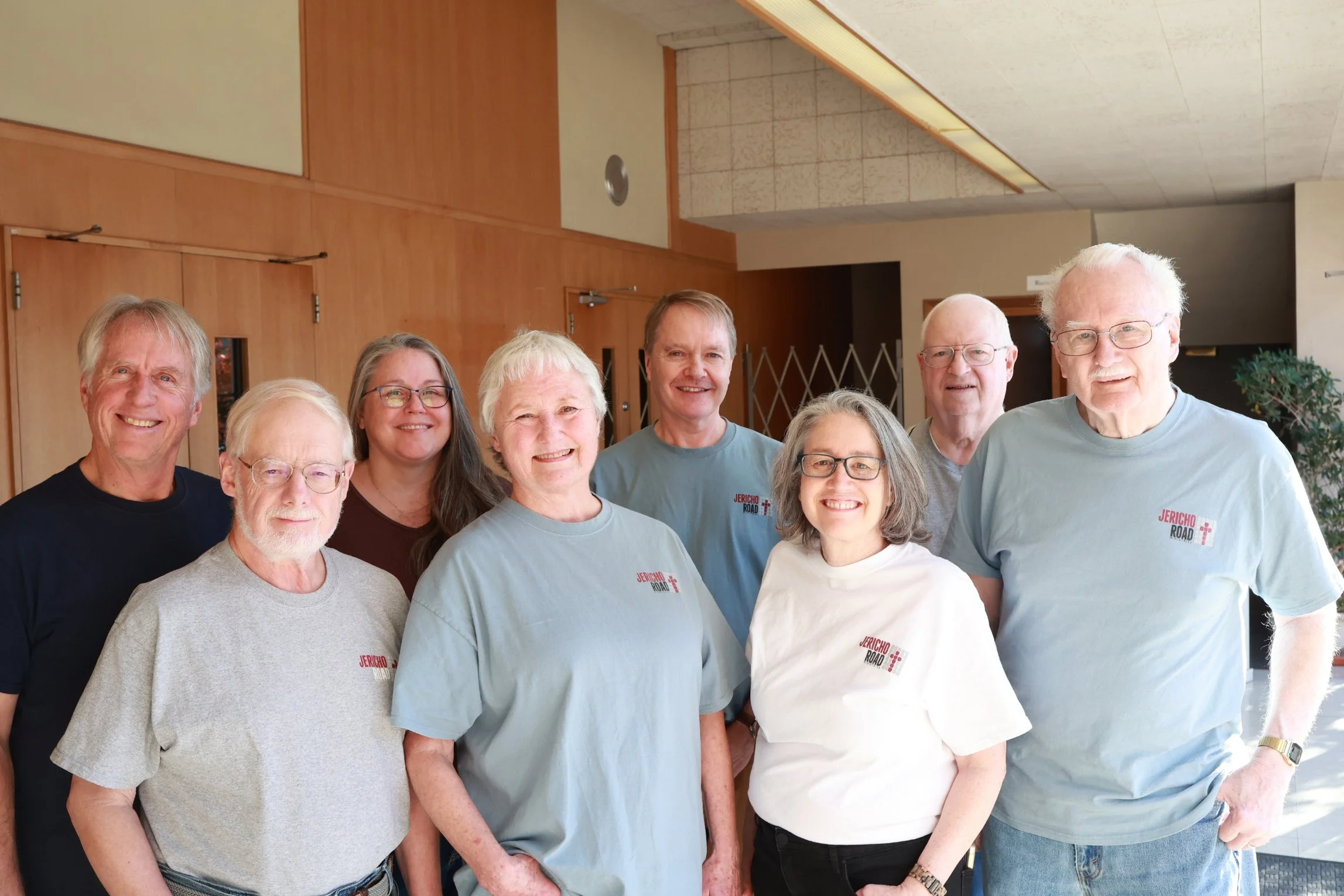 Group of eight smiling people who are on Jericho Road Ministries' Advisory Council standing indoors in front of wooden panel walls.