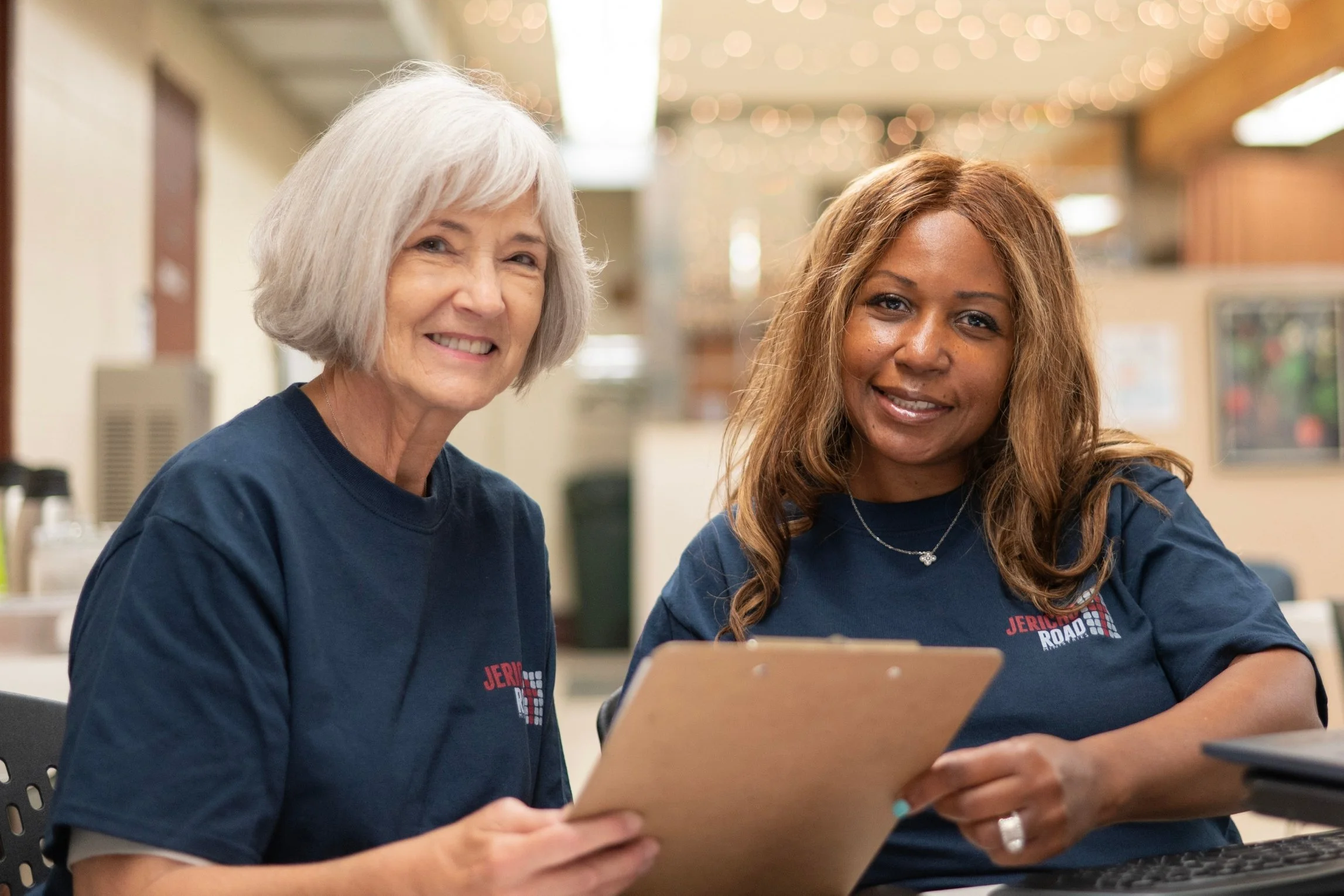 Two women, an older woman and a younger woman, sitting together, smiling, holding a clipboard, inside Jericho Road Ministries' Elliot Park location in Minneapolis.