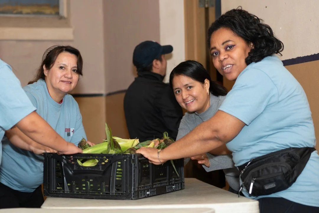 Three women are working together to unpack fresh corn from a black plastic crate. They are smiling and are volunteering at Jericho Road Ministries to serve families experiencing food insecurity.
