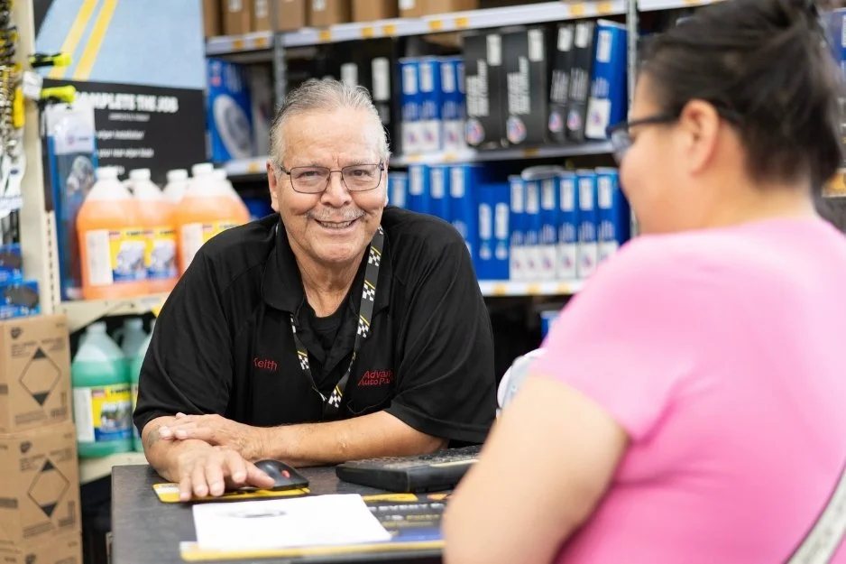 A friendly older man smiling at a woman in pink, standing at a customer service desk inside an auto parts store with various products on shelves in the background.