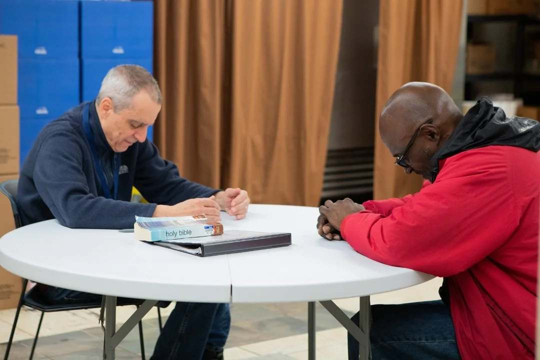 Two men sitting at a round table with their heads bowed, praying. One man has a Bible in front of him, and the other has a binder. The background shows shelves and wooden panels.