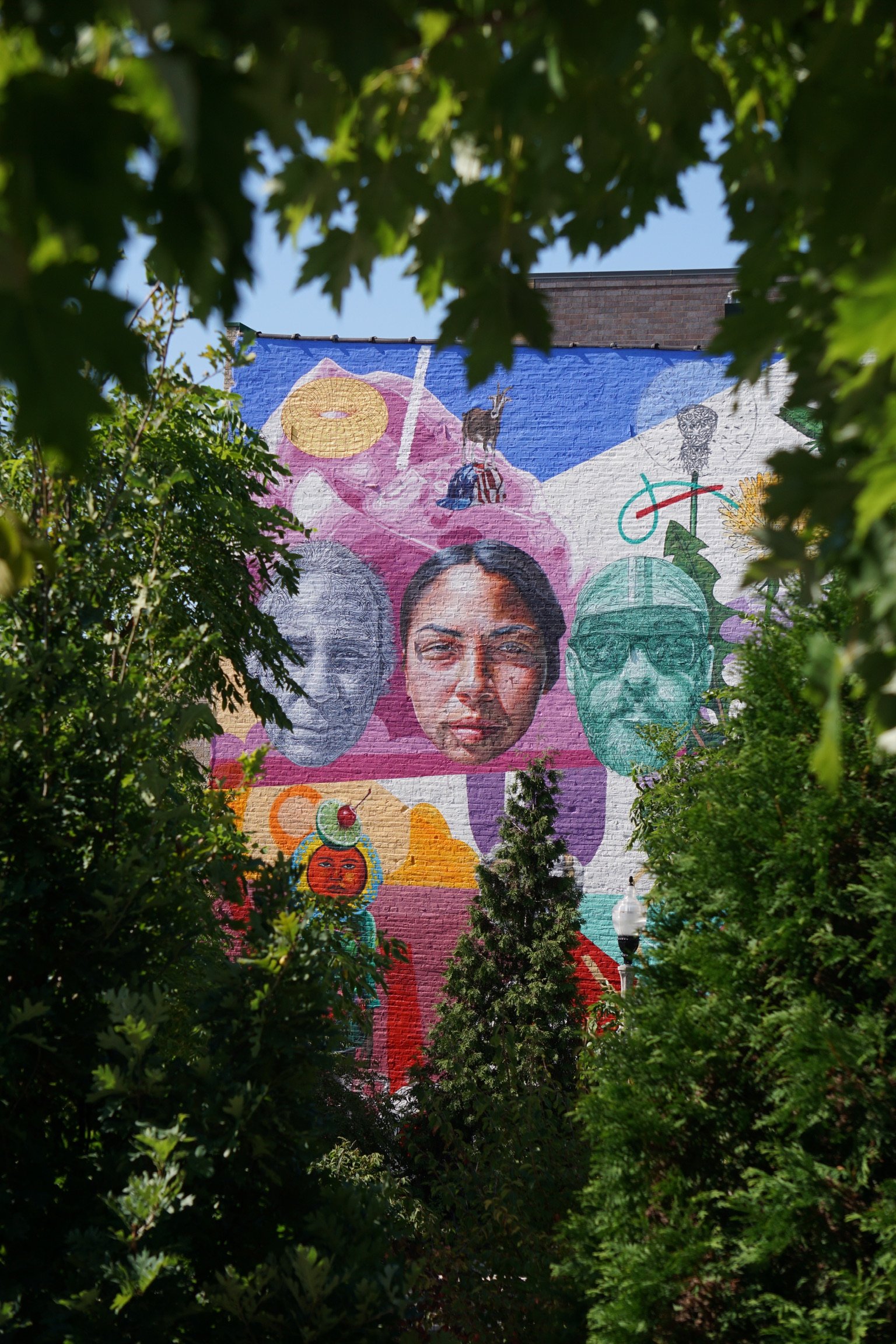 Colorful mural featuring diverse faces, abstract art, and vibrant illustrations, partially framed by green trees.
