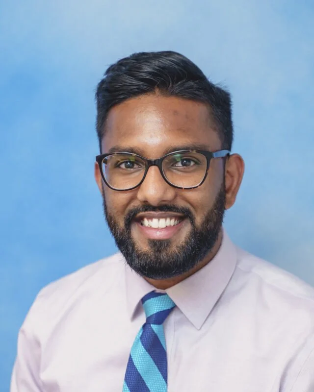 A smiling man with glasses, a beard, and short dark hair, wearing a light purple dress shirt and a blue striped tie, posing against a blue background.