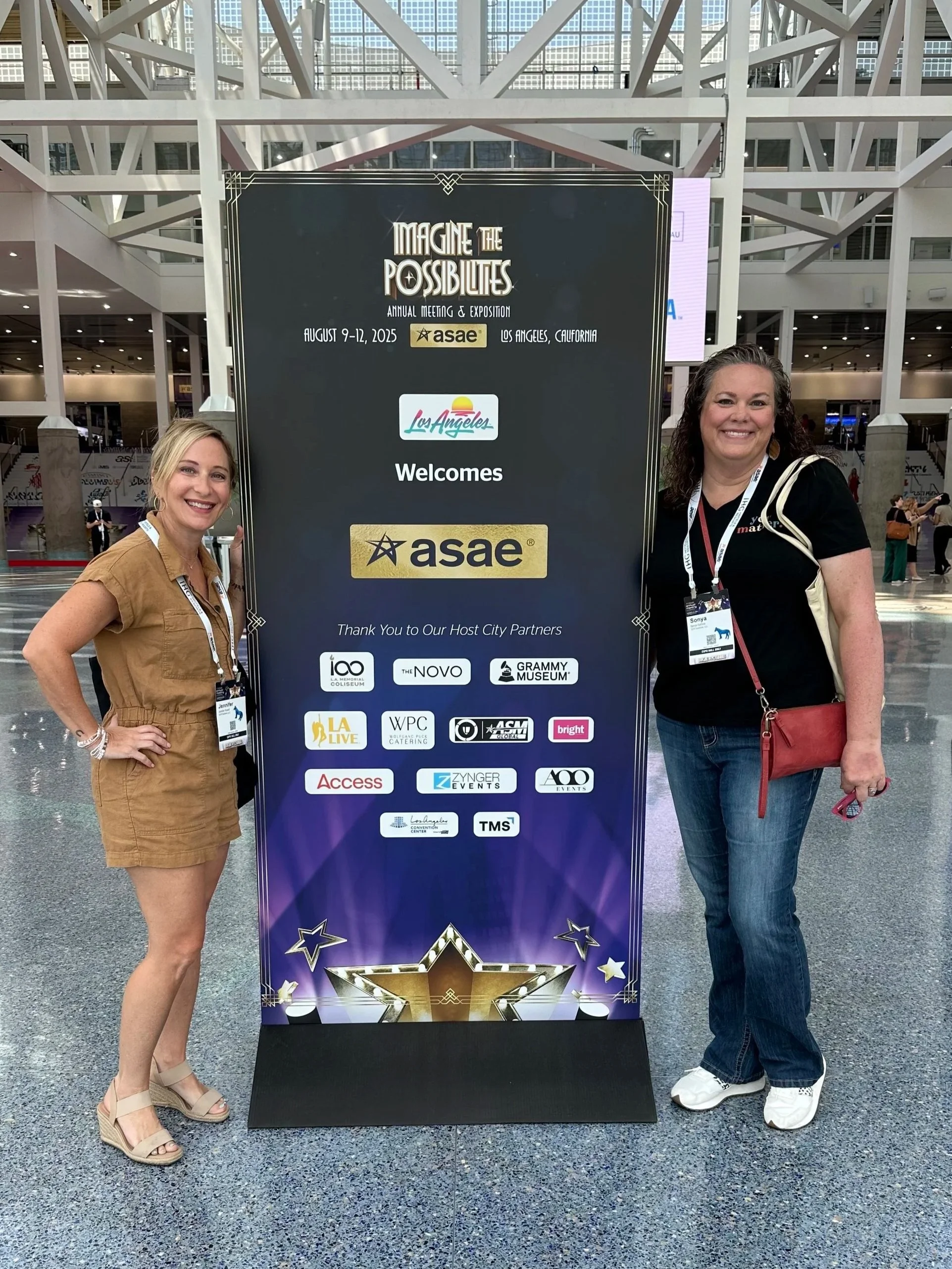 Two women standing next to a large conference sign at an indoor convention center, smiling for the camera.