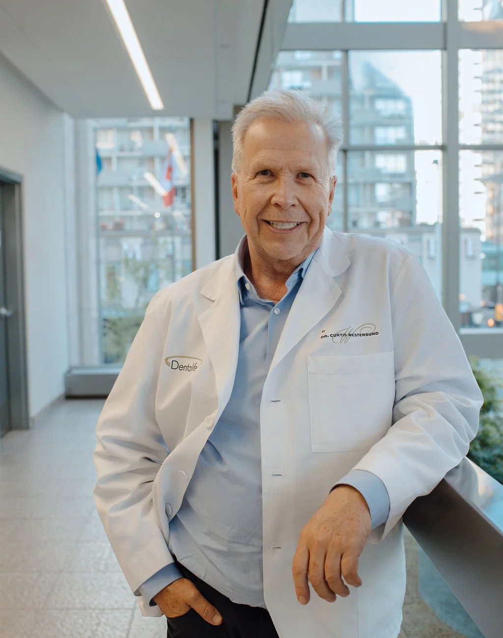 Smiling male doctor in white coat standing in a modern clinic with large windows and city view.